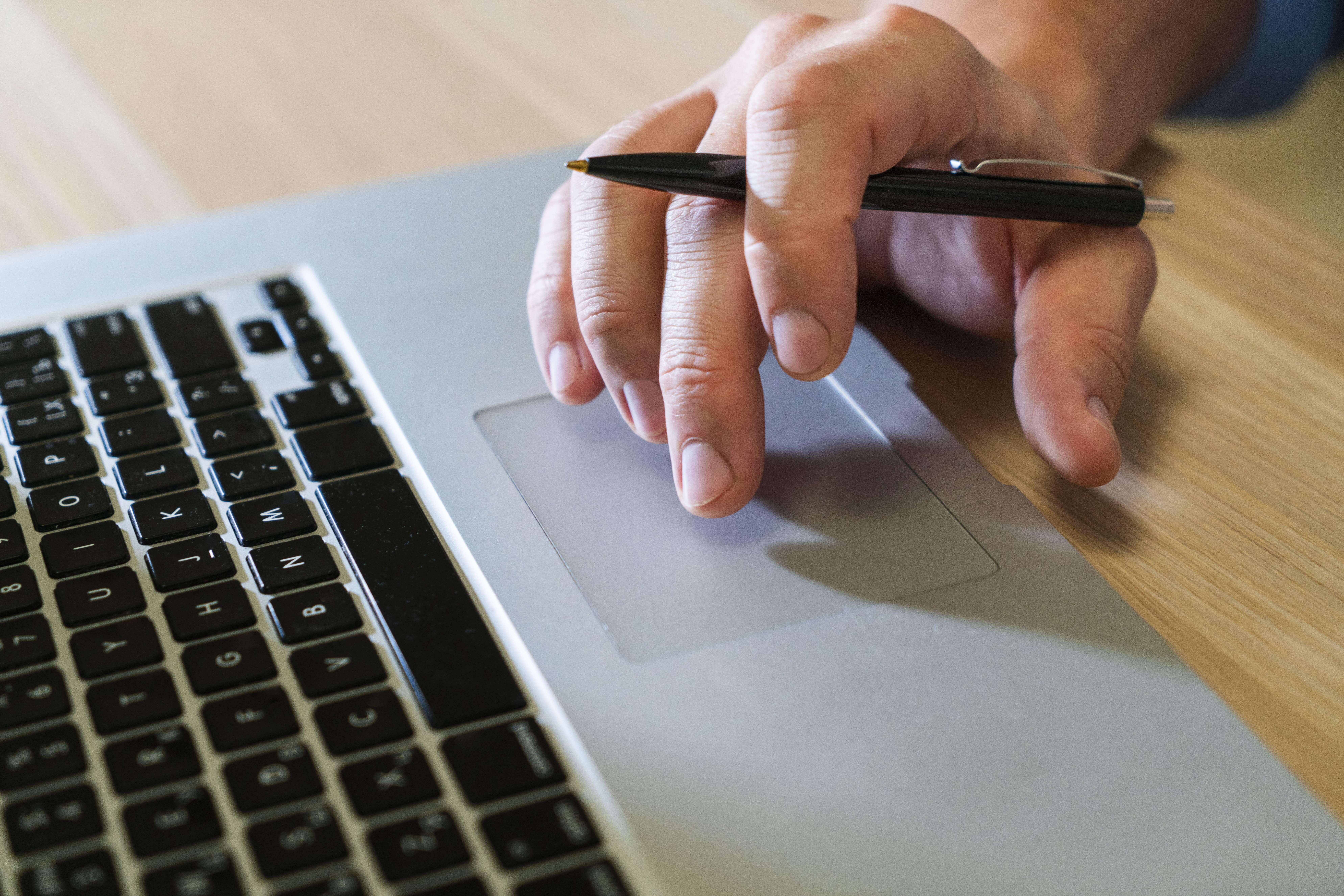 Male hand with pen in it, scrolling on a laptop